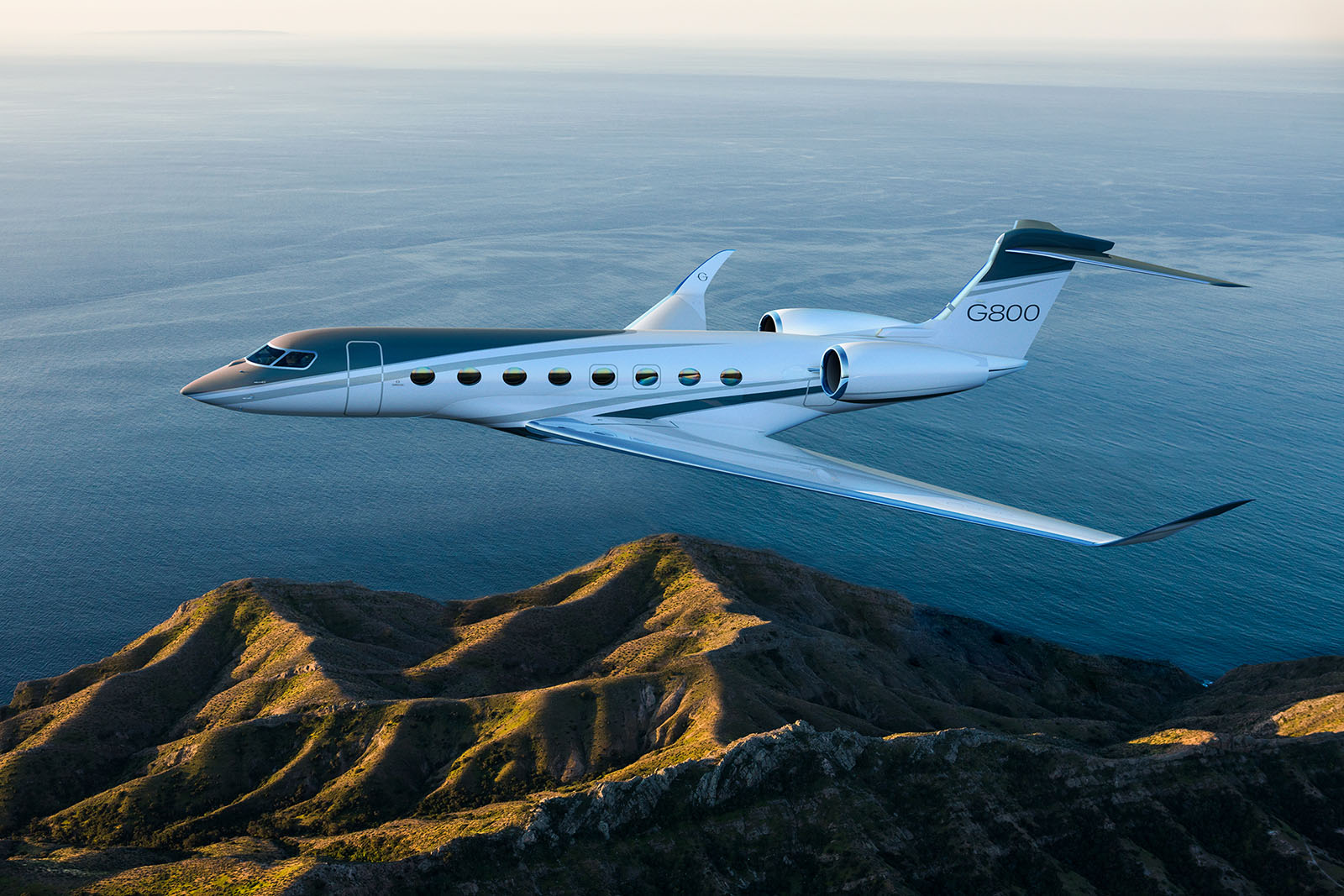 Gulfstream G800 business jet flying over coastal mountains at high altitude during sunset.